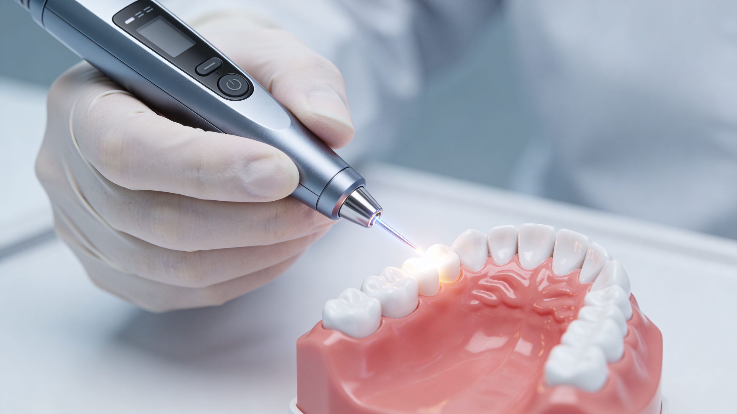 A gloved hand holds a dental laser tool over a model of human teeth and gums.