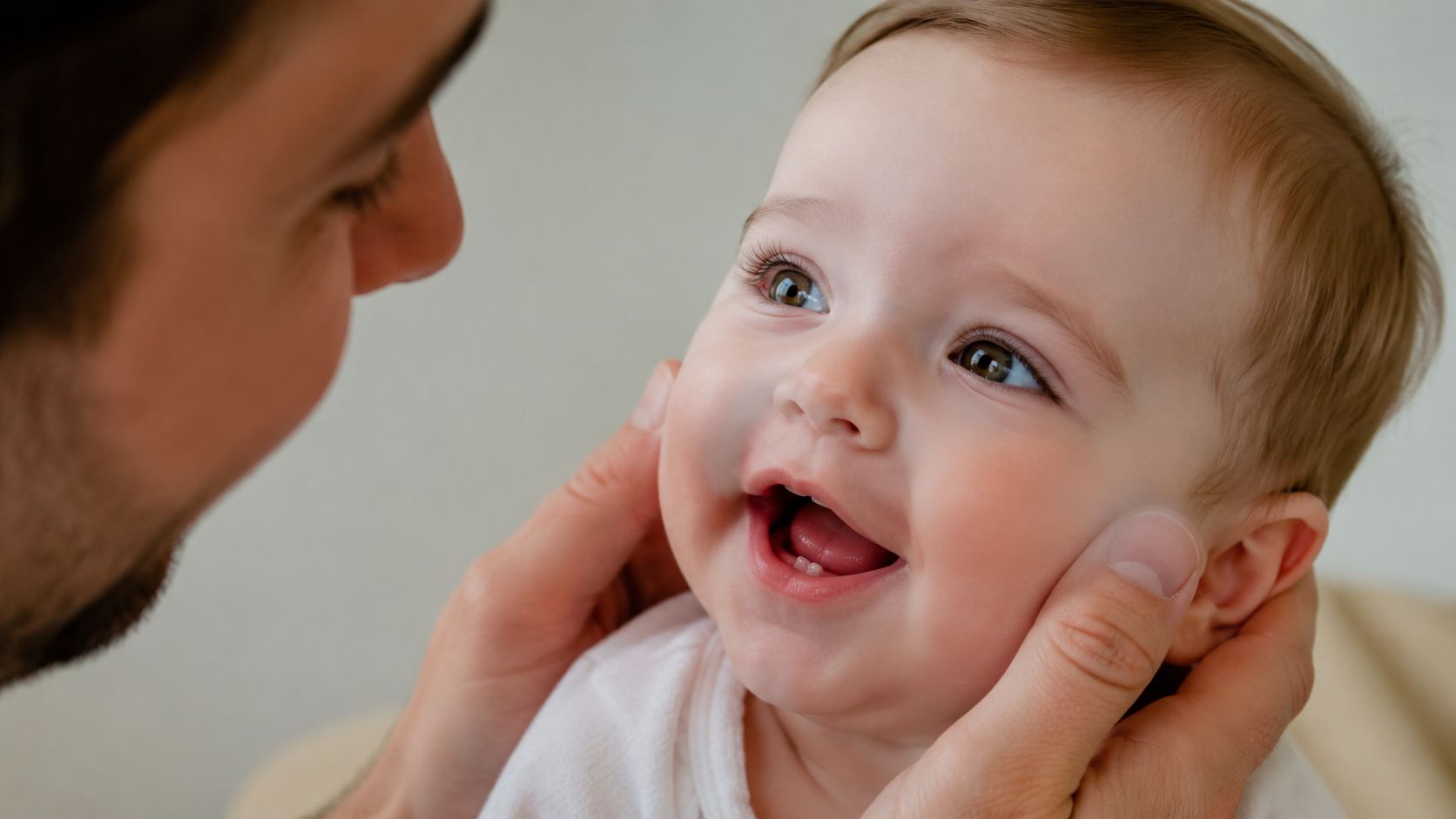 A close-up view of a father gently holding his smiling baby's face to check teeth.