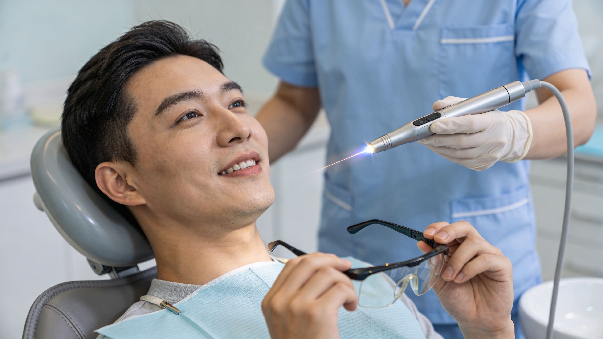 A dentist uses a dental laser tool on a smiling young man in a modern clinical setting.